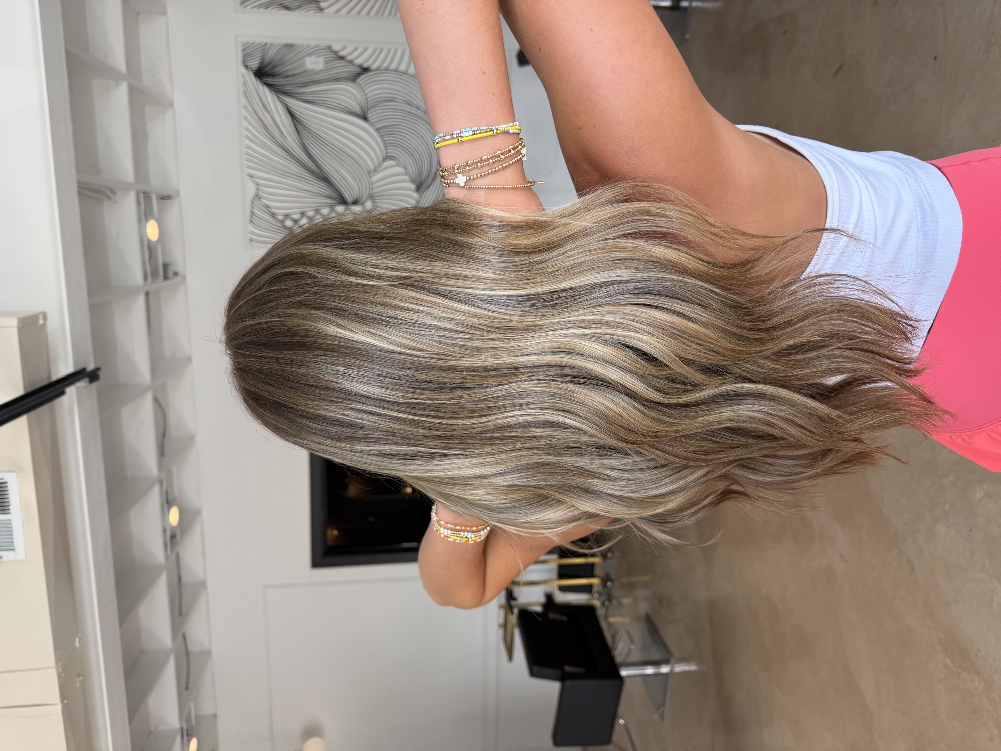 Dimensional blonde balayage seen from behind, hands lifted through the lengths, in a bright modern salon.